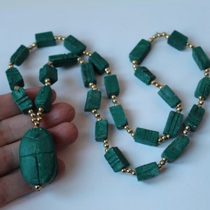 May include: A green malachite necklace with a carved scarab beetle pendant. The necklace is made of rectangular beads with gold spacers.
