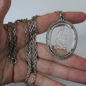 May include: Silver-toned necklace featuring an oval pendant with a clear center. The pendant has a detailed relief of a figure with an animal. The chain is a classic link style.