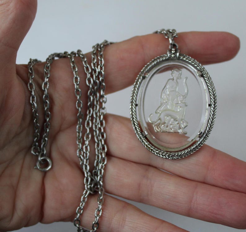 May include: Silver-toned necklace featuring an oval pendant with a clear center. The pendant has a detailed relief of a figure with an animal. The chain is a classic link style.
