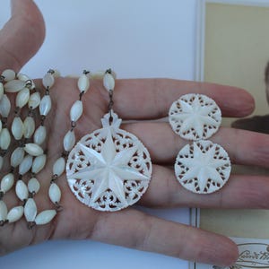 Puede incluir: Un conjunto de joyas vintage con collar, colgante y pendientes. El conjunto está hecho de un material de color claro, posiblemente nácar, con un diseño de estrella. Una fotografía vintage está en el fondo.