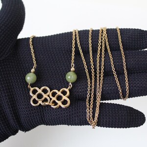 May include: A gold-toned necklace featuring a delicate chain, two smooth round green beads, and a central pendant composed of two interlocking open-work flower shapes. The metal links are thin and uniform, connecting the beads to the floral charm.