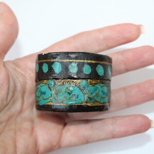 May include: A small, round, black box with a turquoise and gold design. The box has a decorative pattern of dots and swirls.