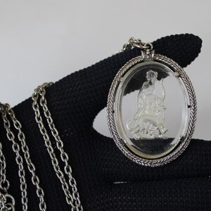 May include: Silver-toned necklace featuring an oval pendant with a clear center. The pendant has a detailed relief of a figure riding an animal. The chain is made of small, linked rings.