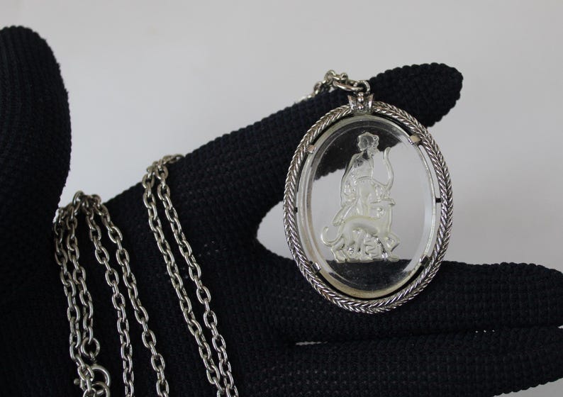 May include: Silver-toned necklace featuring an oval pendant with a clear center. The pendant has a detailed relief of a figure riding an animal. The chain is made of small, linked rings.