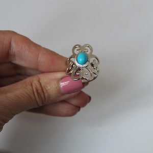 May include: A silver ring with an openwork design featuring a central, round, turquoise-colored stone. The ring's decorative elements create a floral or ornamental pattern. The ring is shown against a plain white background.