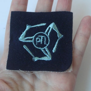 May include: A blue fabric patch with a white embroidered design. The design features a circle with the letters "ATM" inside and three stylized arms extending from the circle.
