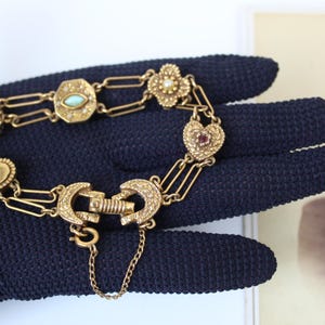 May include: A gold-toned link bracelet featuring a series of decorative charms including a crescent moon, a pearl-set oval, an octagonal piece with a central stone, a clover with a pearl, and a heart with a small red stone, secured by a clasp.