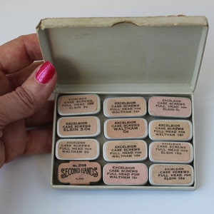 May include: A vintage tin box filled with small tins of watch case screws. The tins are labeled with the brand "Excelsior" and the sizes of the screws. The box is labeled "Second Hands" and has the number 2148 printed on it.