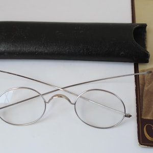 May include: Vintage round eyeglasses with thin silver frames and clear lenses, next to a black leather case. A sepia-toned photograph of a man with a beard is in the background. The glasses have a classic, antique aesthetic.