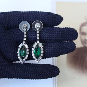 May include: A pair of elegant drop earrings featuring emerald green marquise-cut stones surrounded by clear, sparkling crystals. The earrings have a post backing and are displayed against a dark blue background.