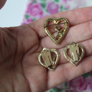 May include: A set of three gold-tone heart-shaped jewellery pieces. The brooch features a delicate floral design and a pin closure. The earrings are clip-on style.