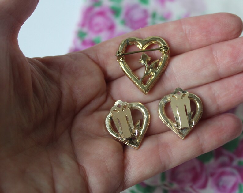 May include: A set of three gold-tone heart-shaped jewellery pieces. The brooch features a delicate floral design and a pin closure. The earrings are clip-on style.