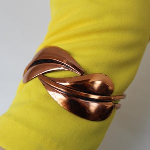 Puede incluir: Un brazalete de tono cobre con un diseño de hoja. El brazalete está sobre un fondo de tela amarilla.