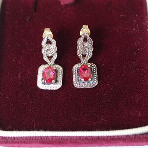 May include: A pair of drop earrings featuring oval-cut red stones set in rectangular frames bordered by small clear stones. The upper portion of each earring consists of an interlocking link design encrusted with tiny clear stones.