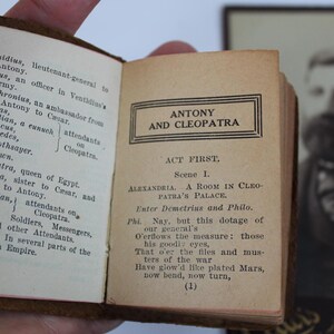 Peut inclure: Un livre ancien ouvert avec le titre "ANTONY AND CLEOPATRA" imprim&eacute; sur la page de droite. La page de gauche &eacute;num&egrave;re les personnages. Un portrait encadr&eacute; est visible en arri&egrave;re-plan.