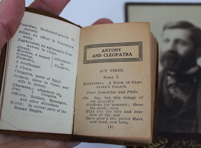 Peut inclure: Un livre ancien ouvert avec le titre "ANTONY AND CLEOPATRA" imprim&eacute; sur la page de droite. La page de gauche &eacute;num&egrave;re les personnages. Un portrait encadr&eacute; est visible en arri&egrave;re-plan.