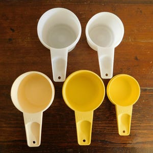 May include: A collection of five measuring cups in various sizes and colors. Two are white, two are yellow, and one is a cream color. Each cup has a handle and a hole for hanging. The cups are arranged on a wooden surface.