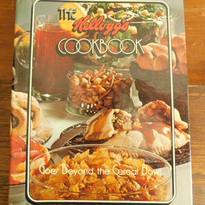 May include: The Kellogg's Cookbook cover features a variety of food items, including a glass of red liquid, a salad, and pastries. The title is in a stylized font with the tagline "Goes Beyond the Cereal Bowl."