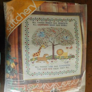 May include: A cross-stitch kit featuring a framed design with a tree, giraffe, lion, and other animals. The text reads "All Things Bright and Beautiful" and "All Creatures Great and Small". The kit is in a clear plastic package.