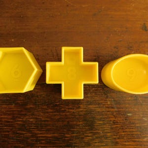 May include: Three yellow plastic molds in distinct shapes: a hexagon with the number 6, a plus sign with the number 8, and an oval with the number 9. Each mold features a raised numeral centered on its base, designed for shaping or casting.