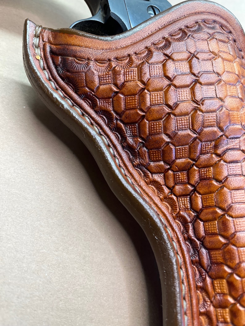 May include: A brown leather holster with a detailed geometric pattern. The holster is designed to hold a firearm.