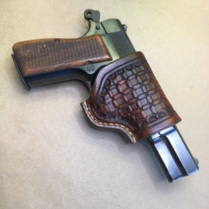 May include: A brown leather holster with a patterned design fits a black handgun with a wooden grip.
