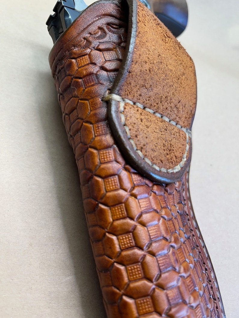 May include: A brown leather holster with a geometric pattern. The holster has a flap that folds over the top and secures with a snap.