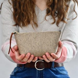 May include: A light beige felt pouch with a brown leather zipper pull. The pouch is held in front of a person wearing a gray sweater with pink and white accents. The pouch has a gold zipper.