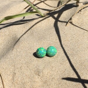 May include: Two green opal earrings with a light green and yellow sparkle. The earrings are on a sandy surface.