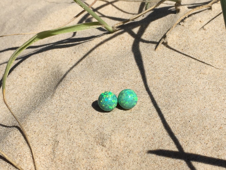 May include: Two green opal earrings with a light green and yellow sparkle. The earrings are on a sandy surface.