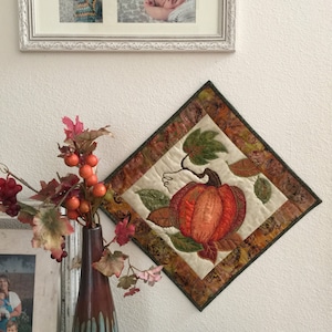 May include: A quilted wall hanging featuring a detailed embroidered pumpkin with leaves and vines in shades of orange, green, and brown. The background is a cream-colored fabric with a paisley pattern.