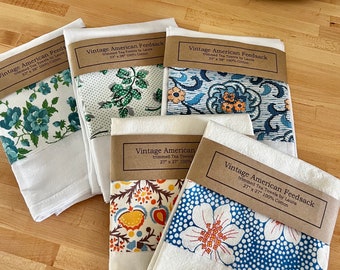May include: Six white tea towels with brown paper labels. The labels have the text "Vintage American Feedsack" and "trimmed Tea Towels by Laura". The towels have different floral patterns in blue, green, orange, and yellow.