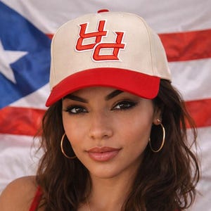 May include: A beige baseball cap with a red brim and a red embroidered design. The cap is worn in front of a Puerto Rican flag. The person is wearing gold hoop earrings and a necklace with a Puerto Rican flag pendant.