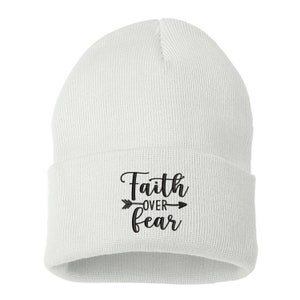 K&ouml;nnte beinhalten: Eine wei&szlig;e Strickm&uuml;tze mit umgeschlagenem Saum. Auf der Vorderseite der M&uuml;tze steht in schwarzer Schrift der Schriftzug "Faith over Fear" mit einem Pfeildesign.