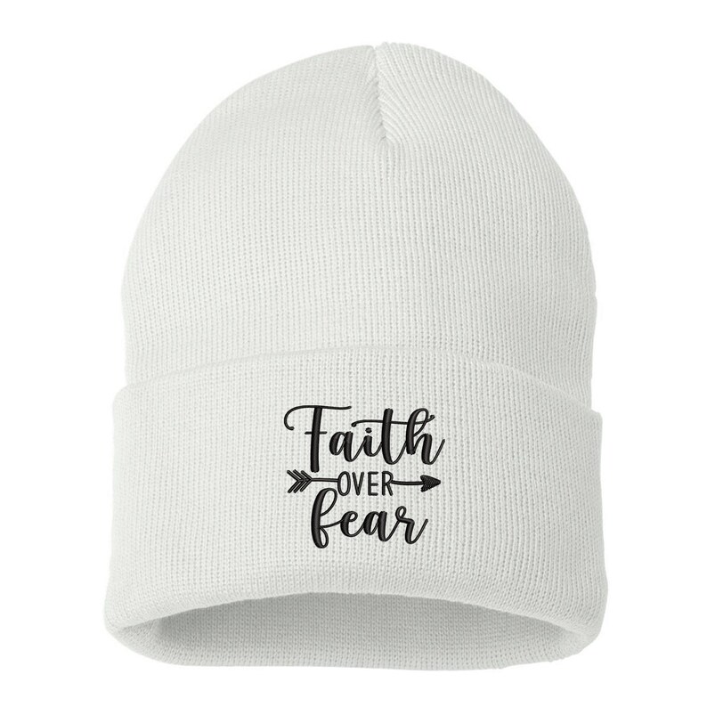 K&ouml;nnte beinhalten: Eine wei&szlig;e Strickm&uuml;tze mit umgeschlagenem Saum. Auf der Vorderseite der M&uuml;tze steht in schwarzer Schrift der Schriftzug "Faith over Fear" mit einem Pfeildesign.