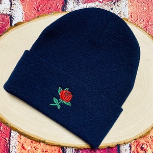 May include: A navy blue beanie with a red rose and green leaves embroidered on the front. The beanie is folded at the brim and is made of a soft, knitted material. The hat is displayed on a light wood surface.