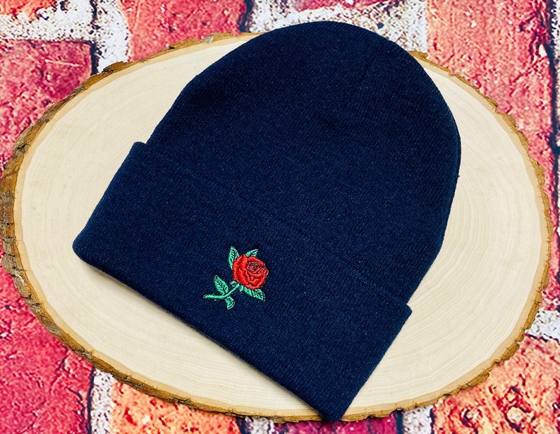 May include: A navy blue beanie with a red rose and green leaves embroidered on the front. The beanie is folded at the brim and is made of a soft, knitted material. The hat is displayed on a light wood surface.