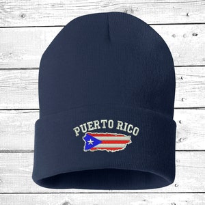 May include: A navy blue beanie with the embroidered text "PUERTO RICO" above an outline of the island in red, white, and blue. The beanie is made of a soft, knitted material.