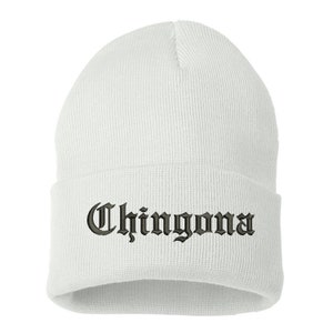 May include: A white beanie hat with the word "Chingona" embroidered in a gothic font. The hat is made of a soft, knitted material and is designed to be worn on the head. The text is in a dark color.