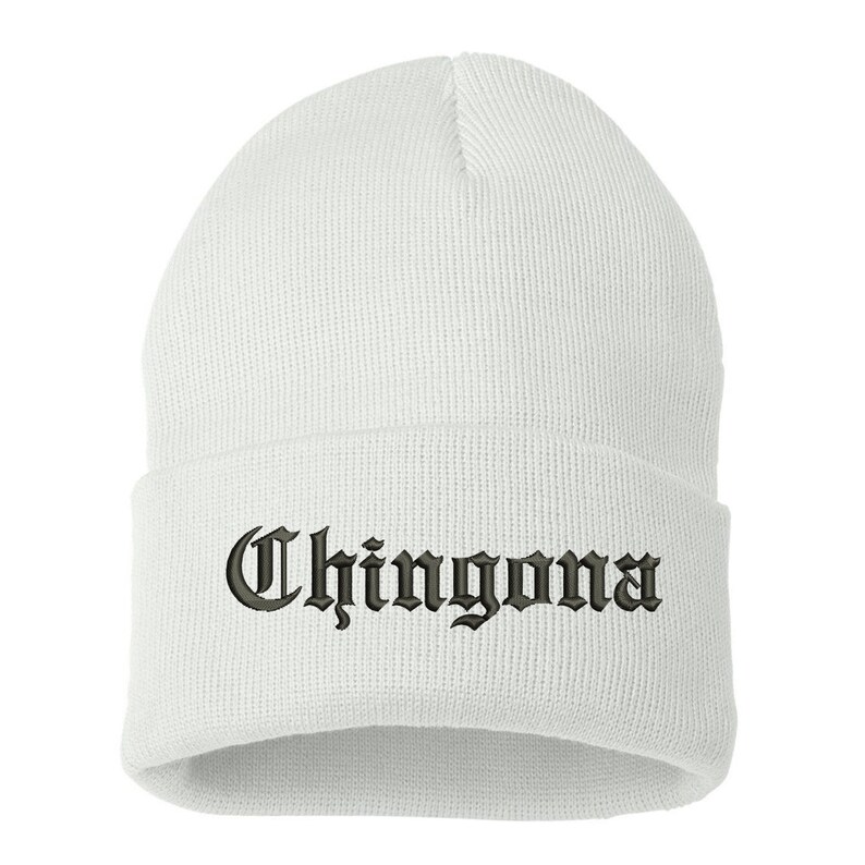 May include: A white beanie hat with the word "Chingona" embroidered in a gothic font. The hat is made of a soft, knitted material and is designed to be worn on the head. The text is in a dark color.