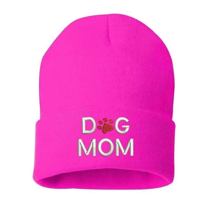 May include: A bright pink beanie hat with the embroidered text "DOG MOM". The text is in white, with a red paw print design between the words "DOG" and "MOM". The hat is made of a soft, knitted material.