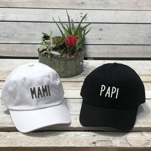May include: Two baseball caps, one white with the word "MAMI" embroidered in black, and one black with the word "PAPI" embroidered in white. The caps are sitting on a wooden surface.