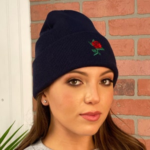 May include: Navy blue beanie hat with a red rose embroidered on the front. The hat is made of a soft, ribbed knit material. The beanie is a stylish accessory for cold weather.