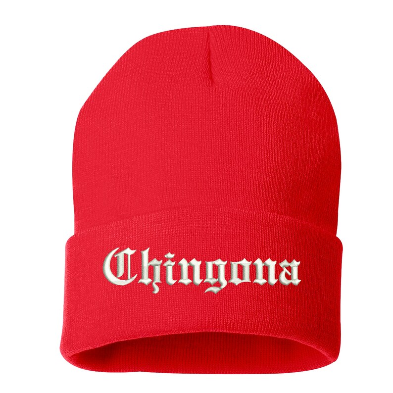 May include: A bright red beanie hat with the word "Chingona" embroidered in white script across the folded brim. The hat is made of a soft, knitted material and is designed to be worn on the head.