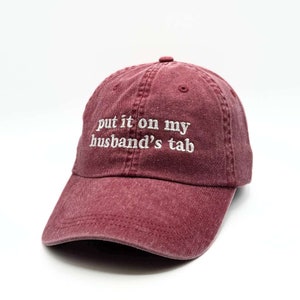May include: A maroon baseball cap with the text "put it on my husband's tab" embroidered in white.