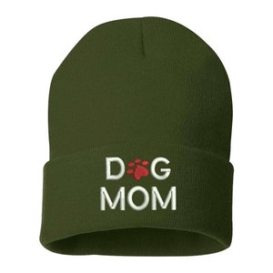 May include: Olive green beanie with the embroidered text "DOG MOM" in white, with a red paw print replacing the "O" in "DOG". The beanie is made of a soft, knitted material.