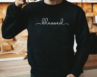 May include: Black sweatshirt with the word "blessed" in a white cursive font across the chest. The sweatshirt is a crew neck style and appears to be made of a soft material. The person in the image is holding a mug.