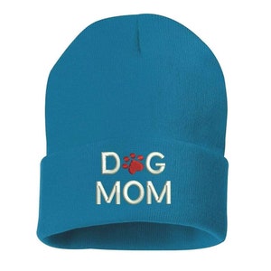 May include: A turquoise beanie hat with the embroidered text "DOG MOM". The word "DOG" is above the word "MOM", with a red paw print in the center. The hat is made of a soft, knitted material.