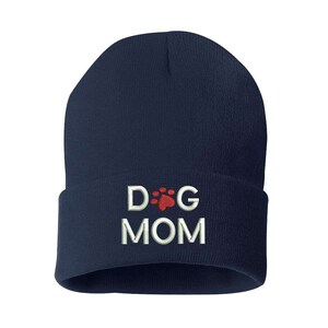 May include: A navy blue beanie hat with a folded brim. The hat features the embroidered text "DOG MOM" in white, with a red paw print design above the word "MOM".