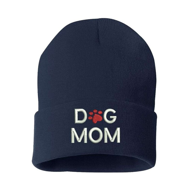 May include: A navy blue beanie hat with a folded brim. The hat features the embroidered text "DOG MOM" in white, with a red paw print design above the word "MOM".